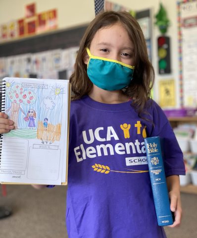 Young girl in school logo t-shirt, wearing a mask but obviously smiling, holds a Bible in one hand and a class coloring assignment in the other. 